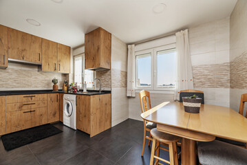 An open kitchen with chestnut wood furniture, black synthetic stone countertops, several windows, large stone floors