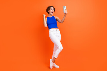 Fototapeta na wymiar Full length photo of delighted pretty person use smart phone raise fist achievement isolated on orange color background