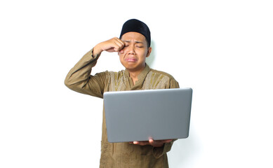 crying asian muslim man holding laptop and rubbing his eye isolated on white background