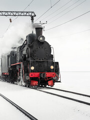 Old steam engine locomotive driving by in a snowy field. Black vintage locomotive pulling a train on a winter day.
