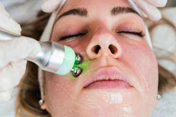 Young woman receiving modern facial low-voltage electrical treatment to stimulate elastin and...