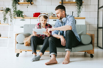 family dad young man with phone and son teenage cute boy with tablet and headphones sit on couch in...