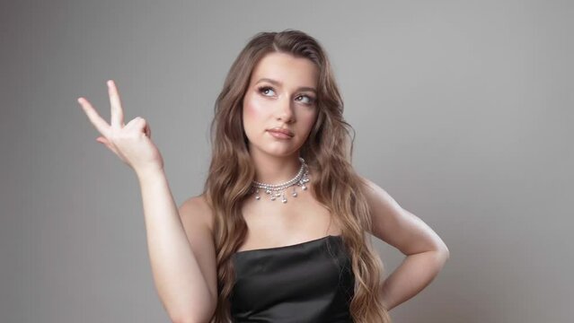 A beautiful young girl with makeup, brown hair in a black spaghetti strap dress mentally counts something on her fingers while posing in the studio on a gray background.