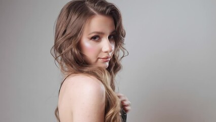 A glamorous girl with brown hair in an evening dress with straps coquettishly shows off her expensive jewelry with diamonds and pearls and poses in the studio correcting her hairstyle.