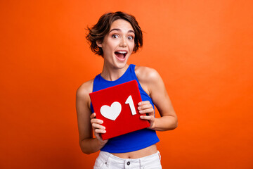 Photo of cheerful impressed lady wear blue singlet open mouth holding heart like card isolated orange color background