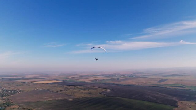Aerial view, stabilized footage along the horizon. A male paraglider flies surrounded by fields and hills. Late fall. Blue-white paraglider wing in sunny weather