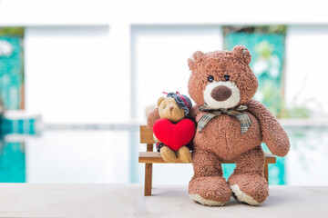 Two teddy bear sitting on wooden bench over blurred swimming pool background, outdoor day light, little teddy bear holding red heart sitting on wooden chair, valentine concept background idea