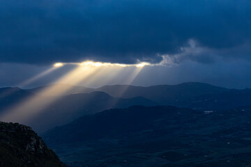 Caccamo, Sicily, Italy Beams of sunlight shine through the clouds illuminating the mountains at sunset.