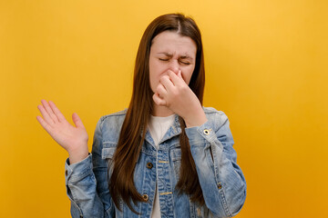 Portrait of sad young woman standing pinching her nose with fingers to hold breath, disgusted by stinky intolerable smell, wearing denim jacket, isolated on yellow studio background. Bad smell concept - obrazy, fototapety, plakaty