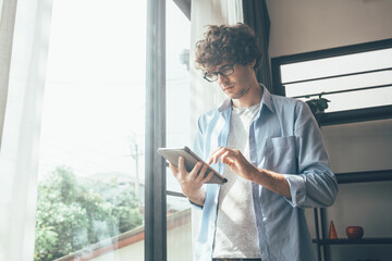 Businessman or creative freelancer man in eyeglasses standing at the window using digital tablet working for customer order or browsing information on the internet at home.