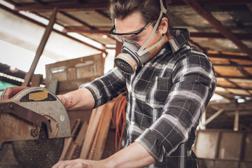 Male carpenter wearing protective mask using electric circular saw cutting wood board at workshop studio