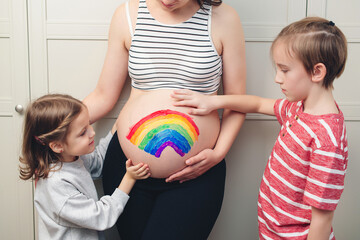Cute boy with sister drawing rainbow on pregnant belly their mother. Baby birth expecting time and belly painting.
