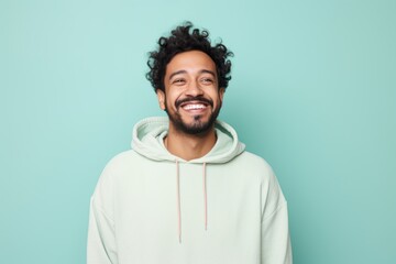 Portrait of a grinning indian man in his 20s wearing a zip-up fleece hoodie against a solid pastel...