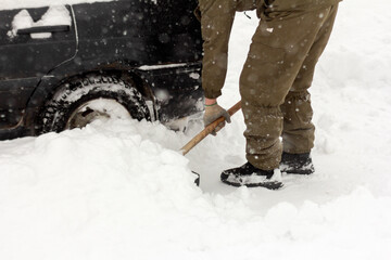 A man with a shovel in his hands removes snow around the car.