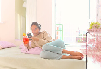 Beautiful young asian woman drinking fruit cocktail and browsing internet on mobile phone at home