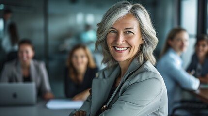 Smiling middle aged beautiful female business person in front of cheerful co-workers in office - obrazy, fototapety, plakaty