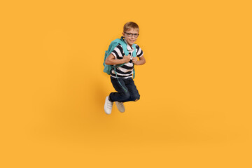 Happy schoolboy in glasses with backpack jumping on orange background