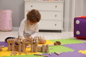 Little boy playing with wooden construction set on puzzle mat in room, space for text. Child's toy