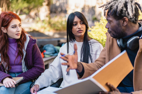Three students engage in a lively discussion with notebooks and papers on a sunny campus day. A sense of collaboration and diversity is evident