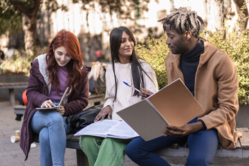 Three students engage in a lively discussion with notebooks and papers on a sunny campus day. A...