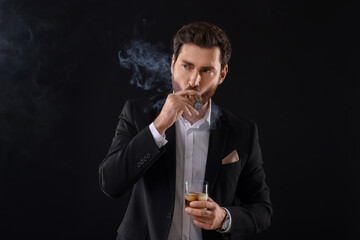 Handsome man in elegant suit with glass of whiskey smoking cigar on black background