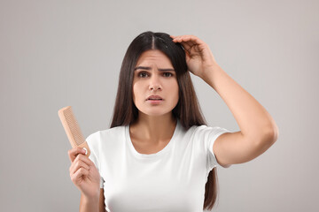 Emotional woman with comb examining her hair and scalp on grey background. Dandruff problem