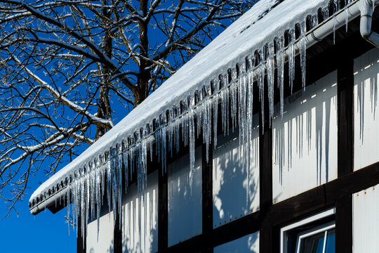 Beautiful icicles hang on the gutter of a historic half-timbered house in Sauerland. Transparent, ephemeral and fragile structures melt and drip in the winter sun. Blue sky and tree in the background.