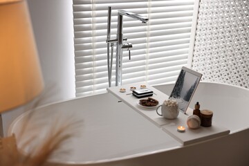 White wooden tray with tablet, spa products and burning candles on bathtub in bathroom