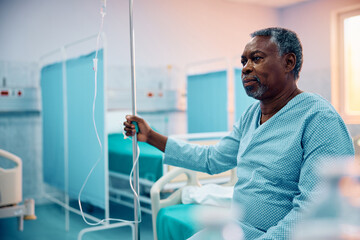 Pensive black senior man with IV drip recovering in hospital ward.