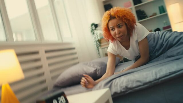 Young African American woman overslept, wakes up looking at alarm clock in panic
