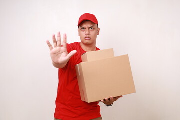 Asian courier man in red holding a cardboard box while showing rejection gesture. Isolated on white