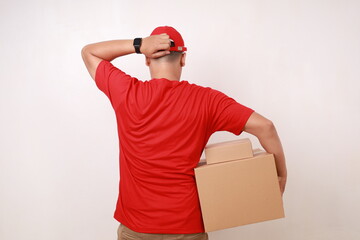 Back view of Confused Asian courier man in red holding head while carrying a cardboard box....