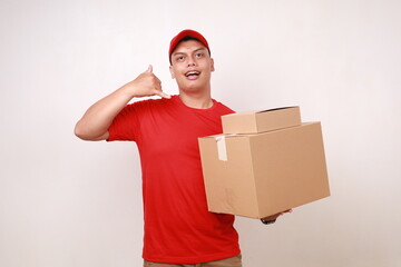 Friendly Asian courier man in red showing phone hand gesture while holding a cardboard box. Isolated on white