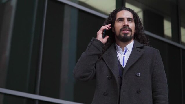 Confident happy mature Latin businessman with long hair looking away while speaking on the mobile phone, outdoors near a modern building