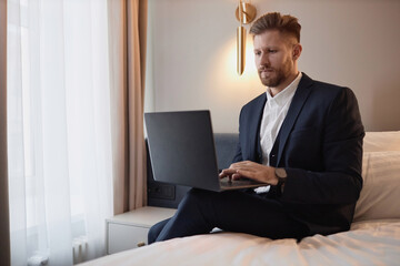 Minimal portrait of successful young businessman using laptop while sitting on bed in hotel room during work trip, copy space 