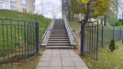 A concrete tile staircase with metal railings leads from the city park through an open gate to the city block. Nearby grassy lawns, trees and buildings. Autumn overcast weather