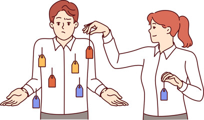 Woman boss tagging confused man employee, for concept of using stereotypes for personnel management. Girl puts price tags on guy shirt, making colleague confused and feel uncomfortable