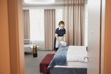 Fototapeta na wymiar Wide angle view at young housekeeper cleaning hotel room and setting up fresh towels on bed, shot through open door, copy space