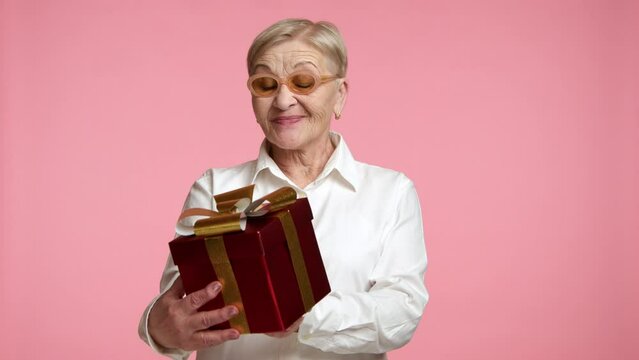 Elegant blonde short-haired senior lady in stylish glasses white shirt looking with admiration at box with present, showing gift to camera, pink background. High quality 4k footage