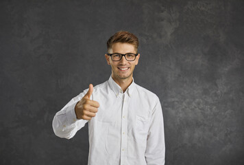 Happy smiling man in glasses giving thumbs up standing isolated on dark grey background. Studio portrait of handsome adult student approving of great idea, showing like gesture or doing okay hand sign