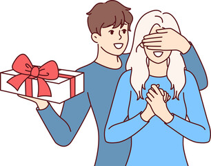 Groom surprises girlfriend standing with gift box and covering girl eyes with hand. Boyfriend in love congratulates happy bride on birthday and Christmas or valentine day on february 14th.