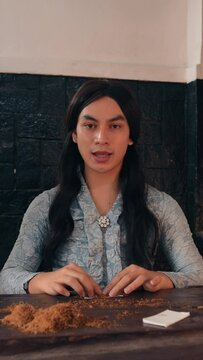 Serious young adult with long hair sitting at a table with a rustic backdrop looking at the camera with a neutral expression., wearing a patterned shirt