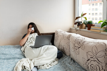 Young sick woman wrapped in a blanket wipes her nose with paper tissues, got sick because she...