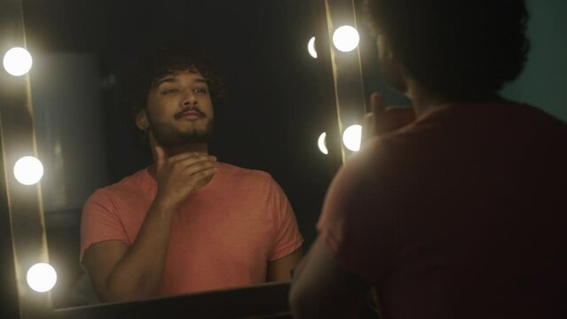 An attractive stylish smiling indian guy wears an earring, in front of the mirror in the dressing room, corrects makeup and hairstyle, prepares for stage or professional photo shoot