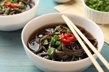Tasty soup with buckwheat noodles (soba), chili pepper, green onion in bowl and chopsticks on light blue wooden table, closeup