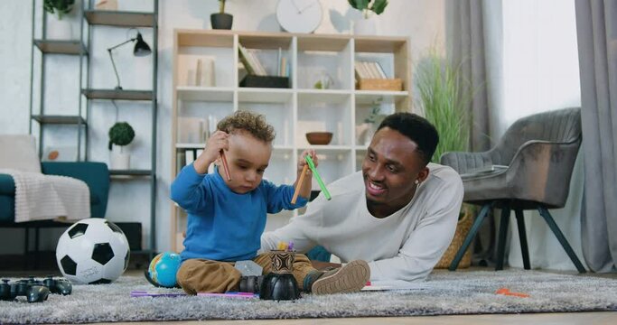 Handsome smiling happy african american man lying on the floor and helping his cute baby boy to gether coloured pens child care concept