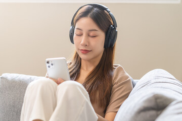 Relaxing asian young woman using smart phone listening to music, enjoy song or watching videos, podcast on mobile with white wireless headphones sitting on sofa, couch at home, chill out and leisure.