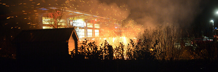 Riesiger Funkenflug aus dem qualmenden Feuer vor der Feuerwache mit dicker Rauchwolke. Scharfer Vordergrund mit Gartenhaus und Hecke in Silhouette. Panorama Bild.