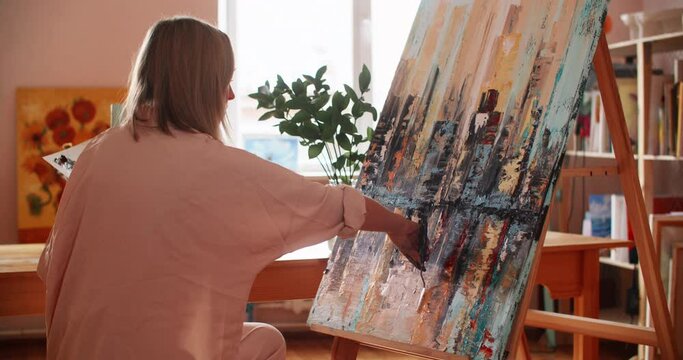 Talented painter painting on canvas with palette knife spatula. Skilled female artist is creating textured art. She is working at workshop on sunny day.