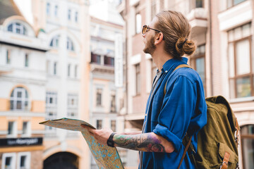 Modern tourist. Handsome young man in casual clothing holding map and looking away while standing outdoors trying to find a direction route in a new unknown city